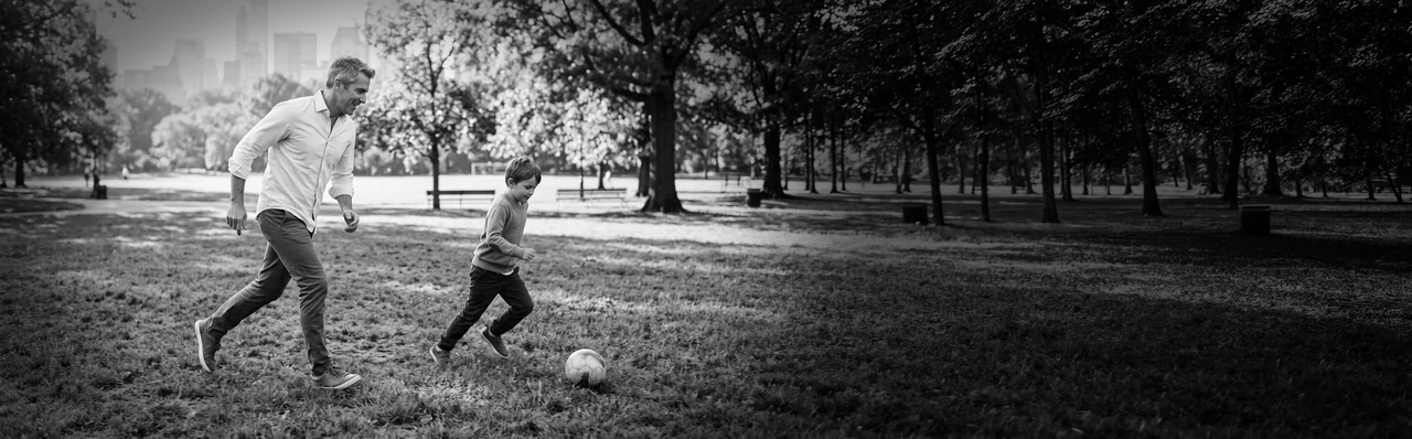 A father and his young son playing football on the grass in a tree-lined park