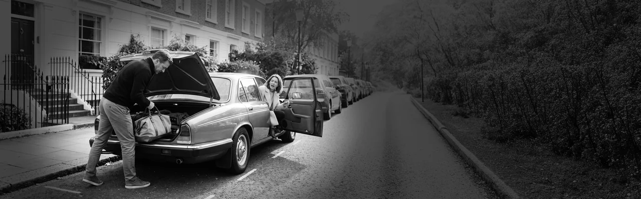 A man loading bags into the boot of a classic car on a terraced street while a woman waits by the open door