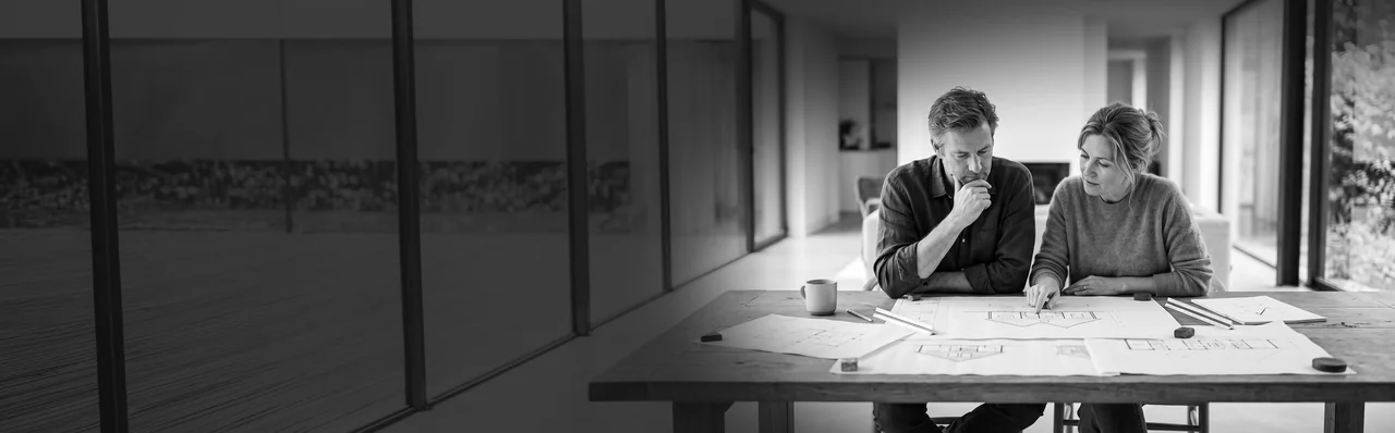 A couple studying drawings spread across a table in a bright room